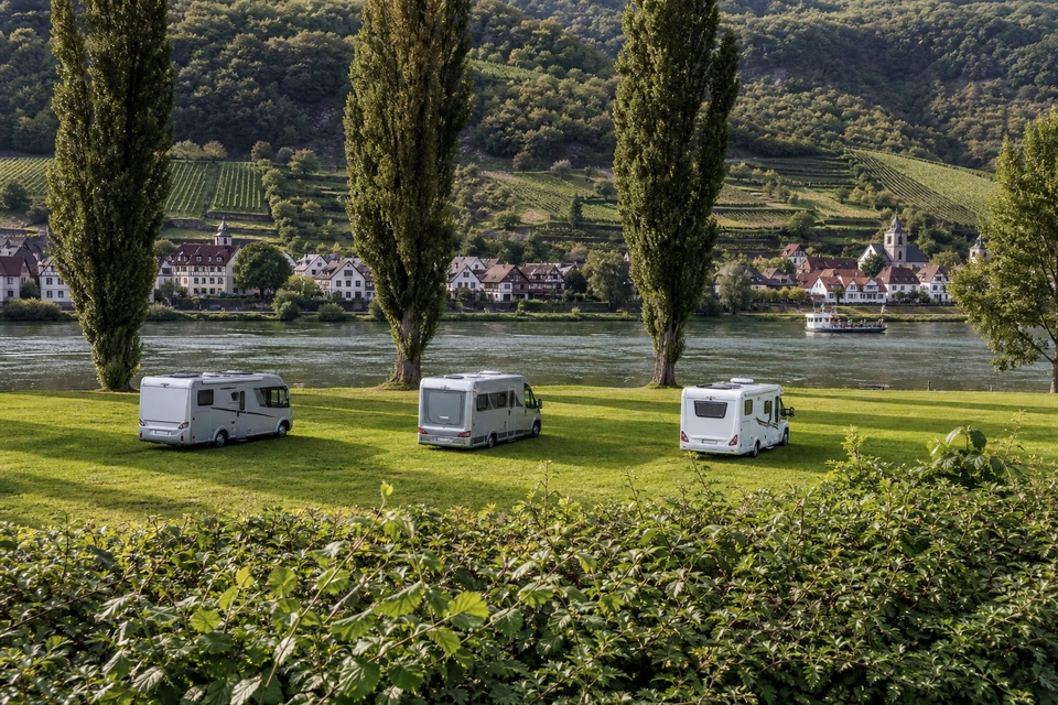 Drei Wohnmobile in gleichmäßigem Abstand auf einer gemähten Flusswiese (mit AI erstelltes Bild)