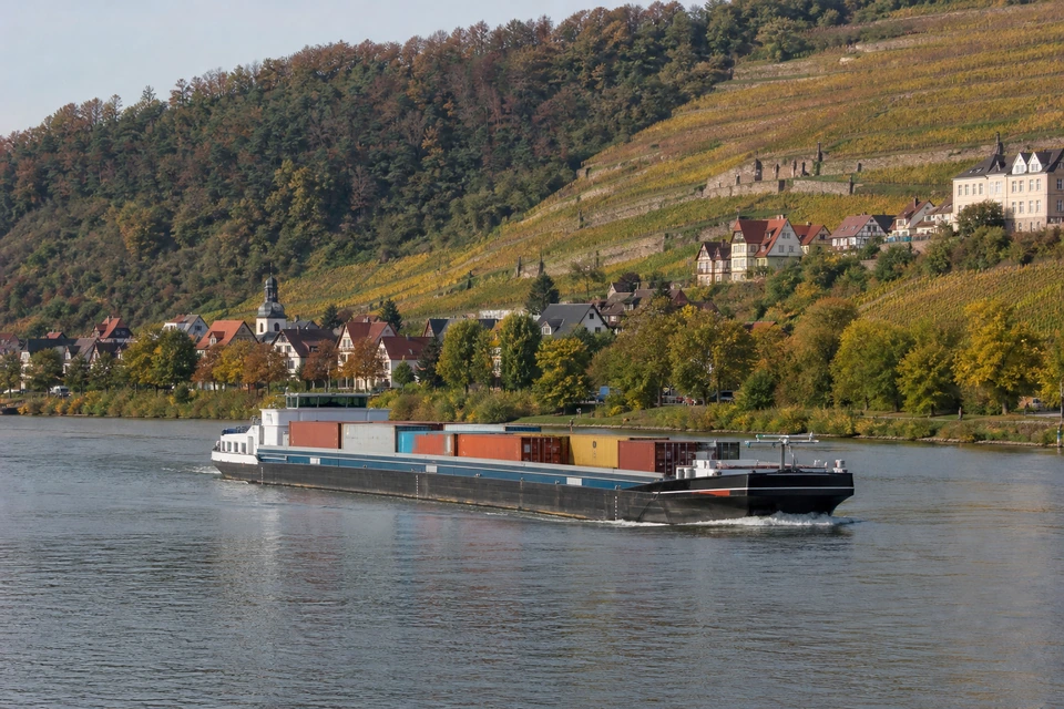 Beladenes Binnenschiff auf einem breiten Fluss, dahinter ein terrassierter Weinberghang (mit AI erstelltes Bild)